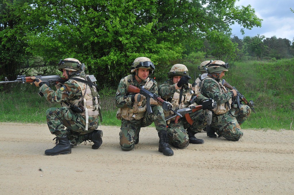 A group of soldiers kneeling in a line on a road with weapons shouldered, trees behind them