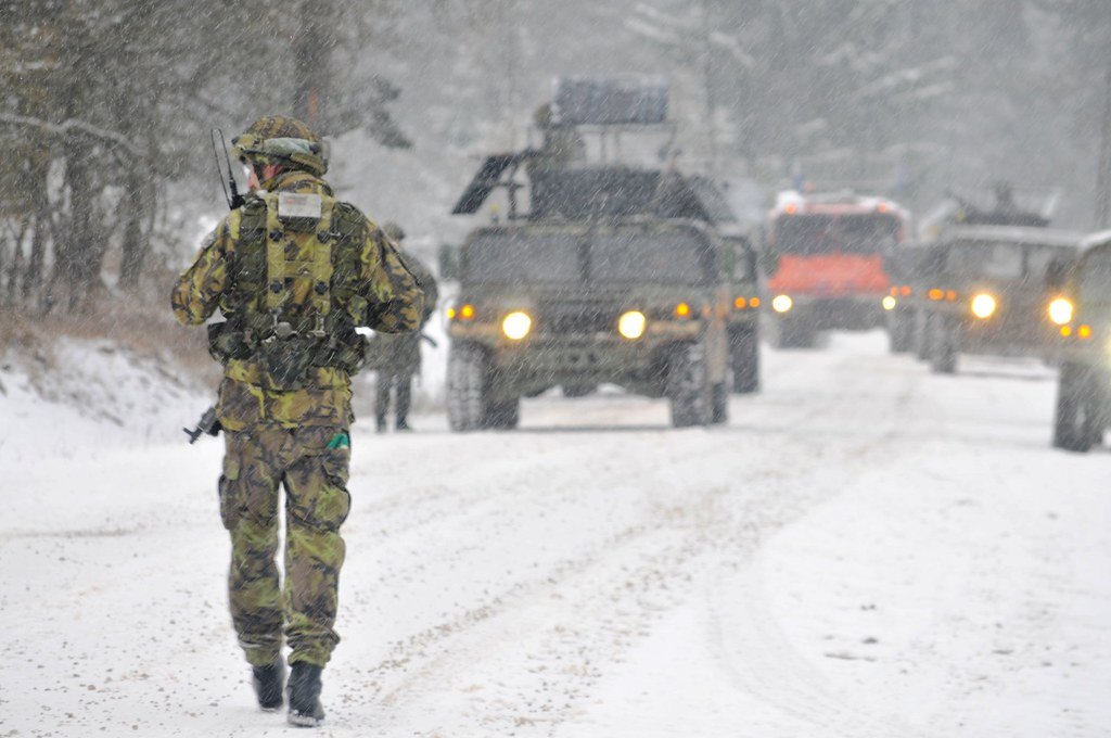 A soldier in winter kit walking along a snow covered track beside a column of military vehicles