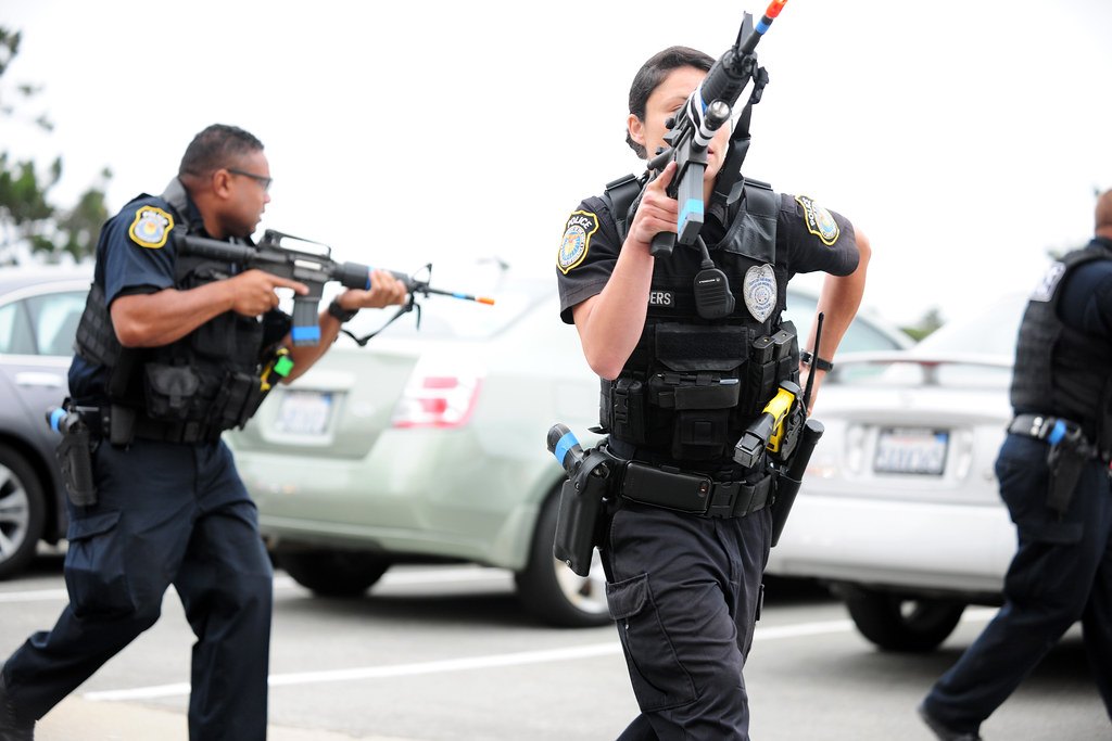 Two uniformed officers with long guns moving between parked cars during a marked training exercise