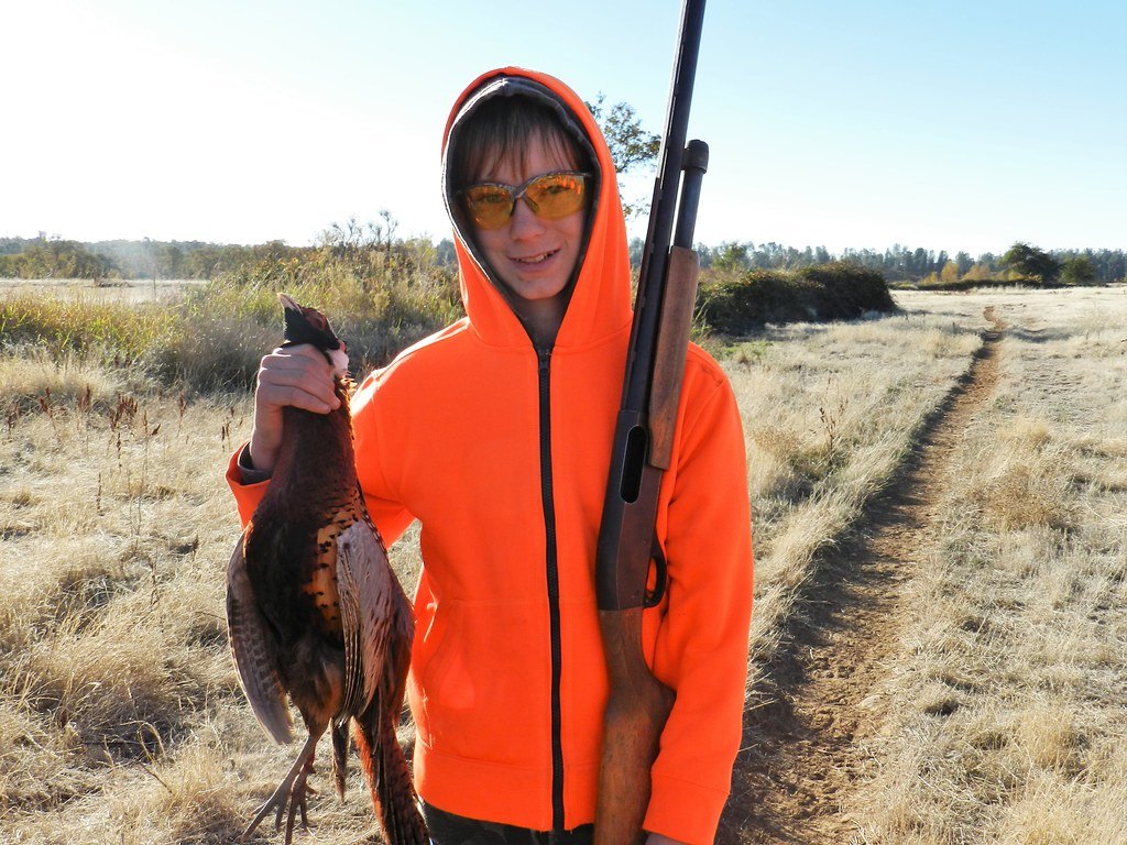 A hunter in an orange jacket holding a pheasant, a shotgun resting over the shoulder, on a grass track