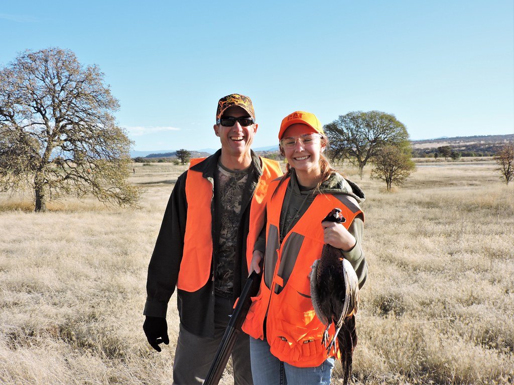 Two hunters in blaze orange standing in a dry field holding a bird they have taken