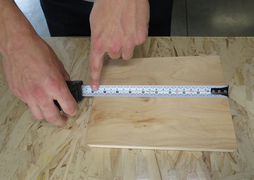 Two hands holding a tape measure across a board and marking a point with a pencil