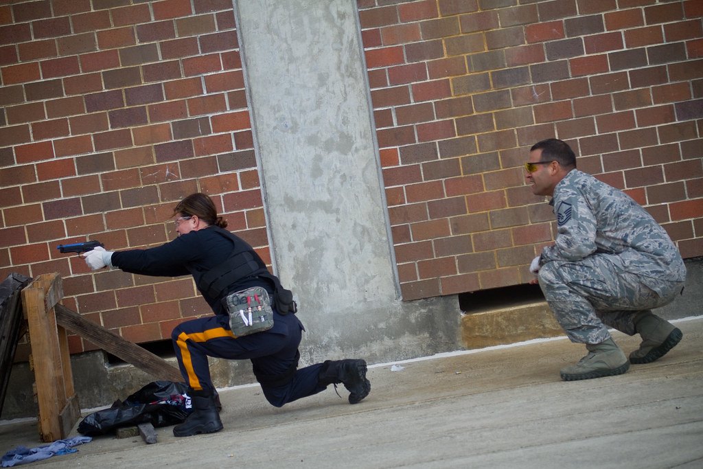 Officers in a training exercise moving alongside a brick building, one kneeling behind cover