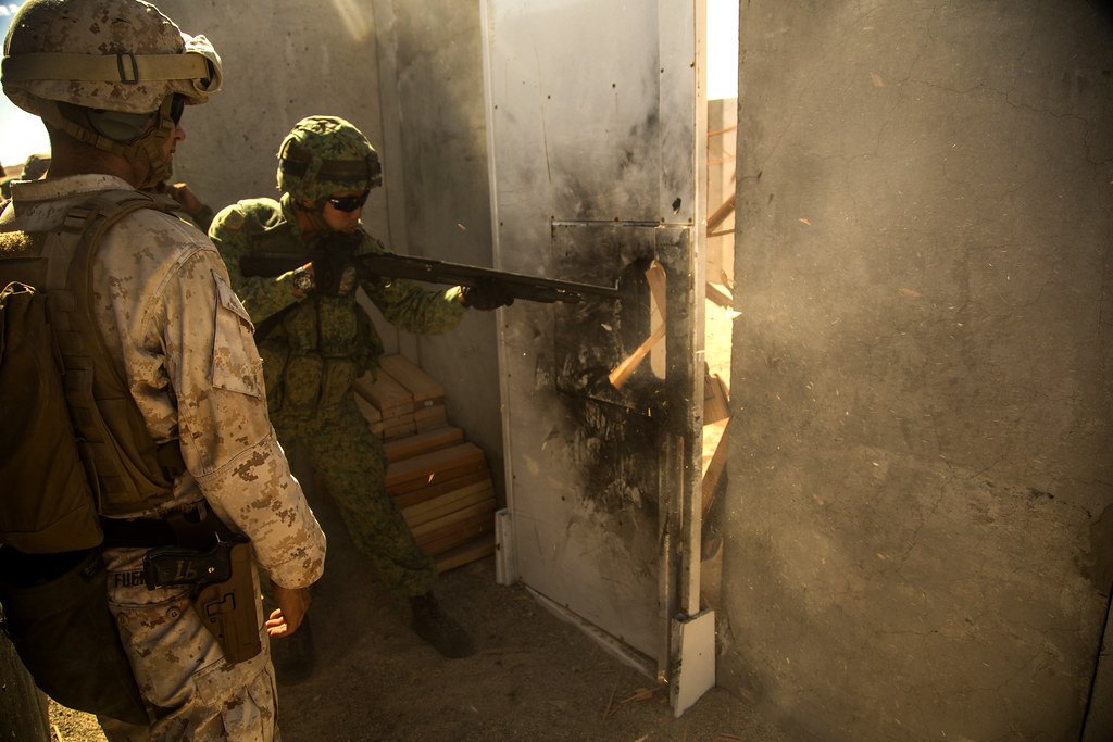 Two service members at a doorway during a training exercise, one working a shotgun against the door edge as another waits behind