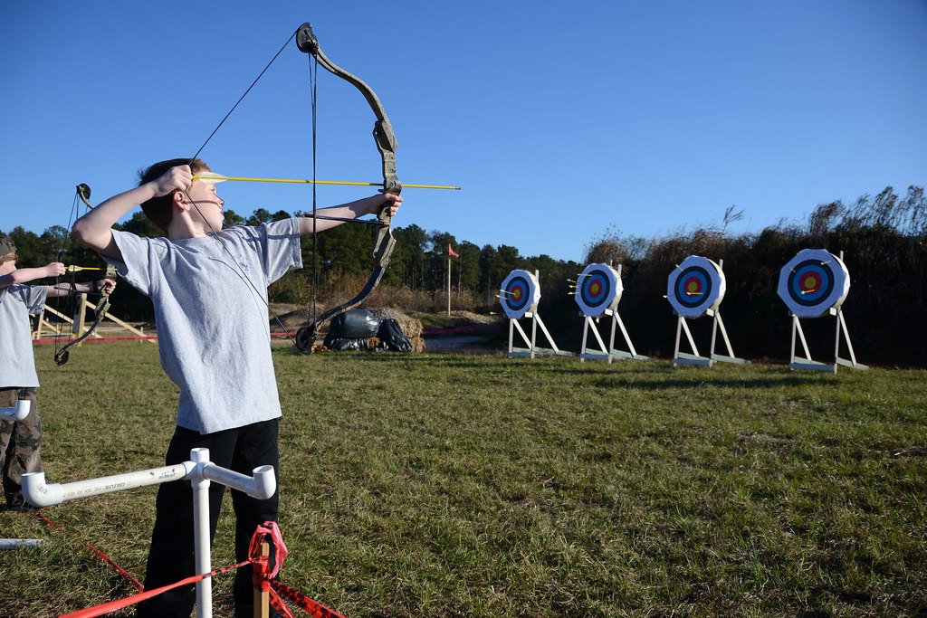 An archer at full draw on an outdoor grass range with four round target faces on stands behind