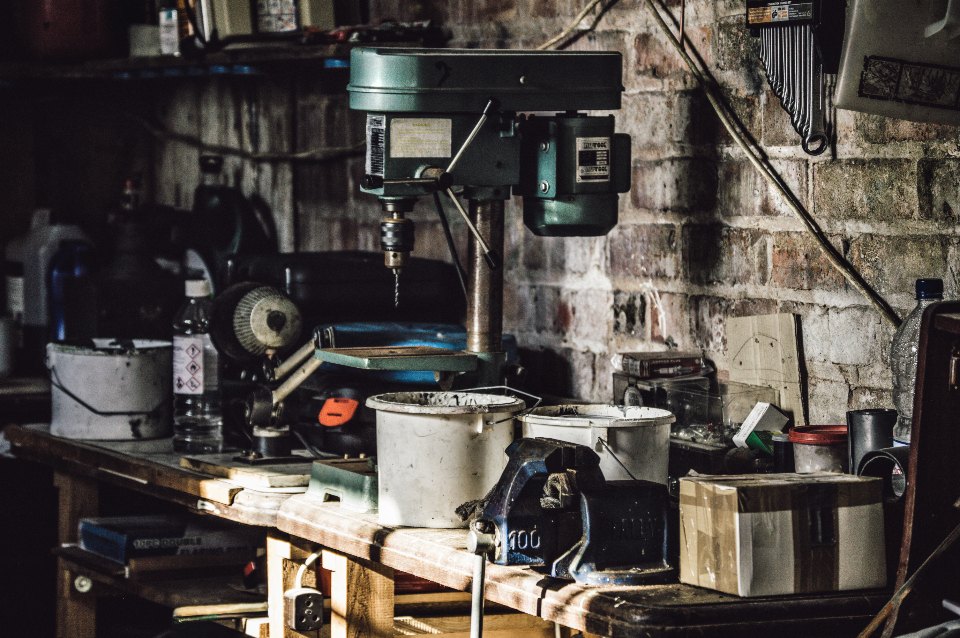 A cluttered garage workbench with a bench drill press, tins, bottles and a vice