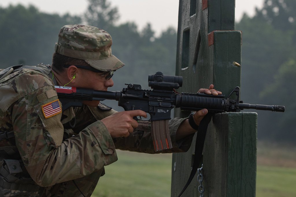 A soldier firing a rifle from behind a wooden barricade on an outdoor range, the sling hanging in a loop beneath the rifle