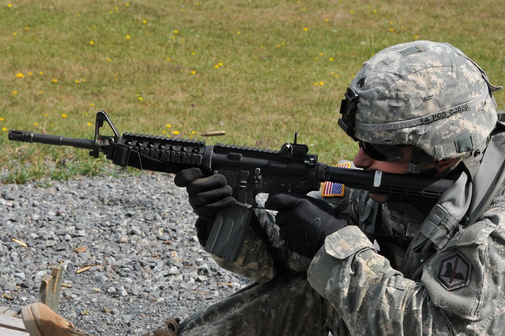 A soldier lying prone on a gravel firing point aiming a rifle, with a sling running along the side of the rifle