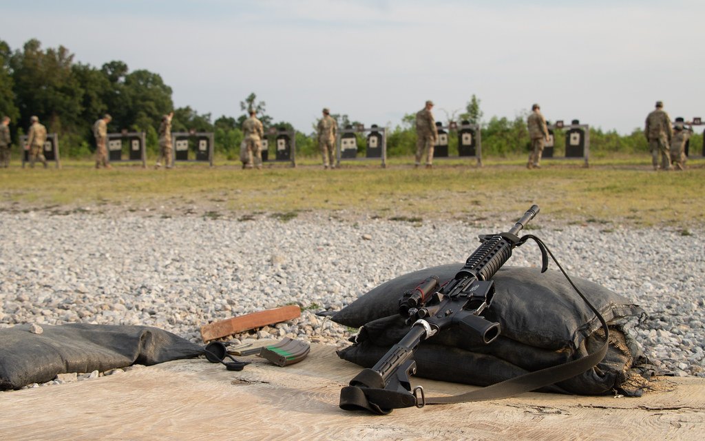A carbine fitted with a sling resting on a sandbag at a gravel firing point, with silhouette targets in the distance