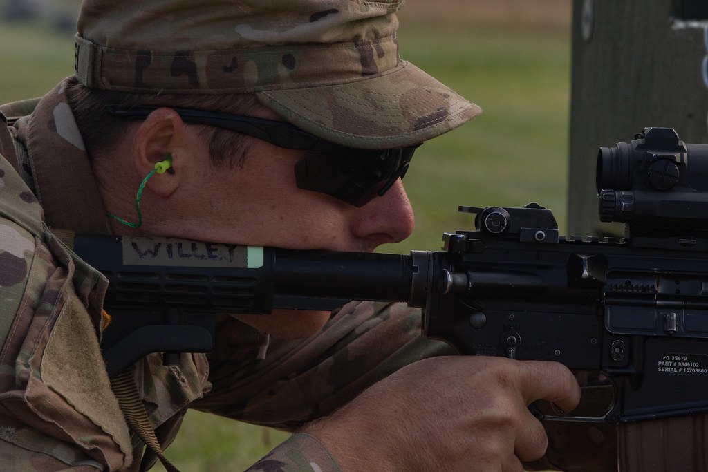 A shooter in profile aiming a rifle fitted with a small optic, wearing sunglasses and foam ear plugs