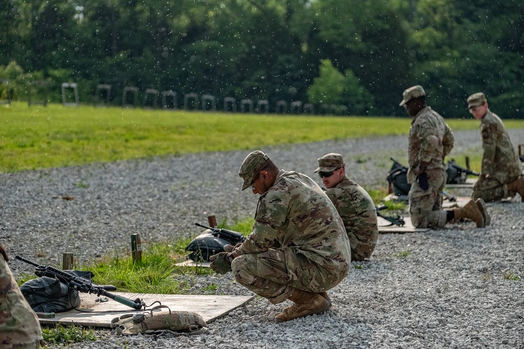 Soldiers kneeling on a gravel firing point beside rifles and equipment on an outdoor range