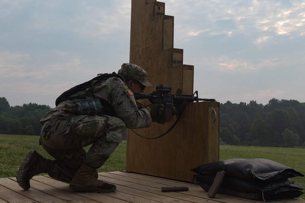 A soldier kneeling behind a wooden barricade aiming a rifle, with sandbags on the platform and open country beyond