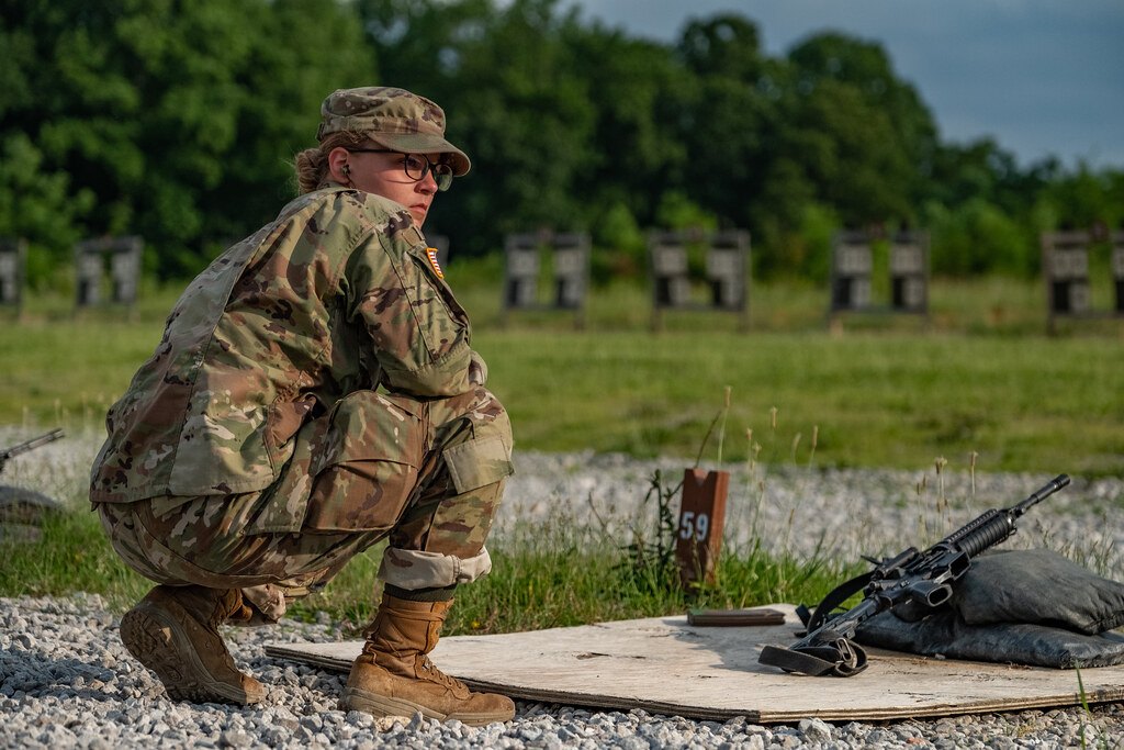 A soldier kneeling on a gravel firing point beside a rifle resting on a sandbag, with target frames in the background