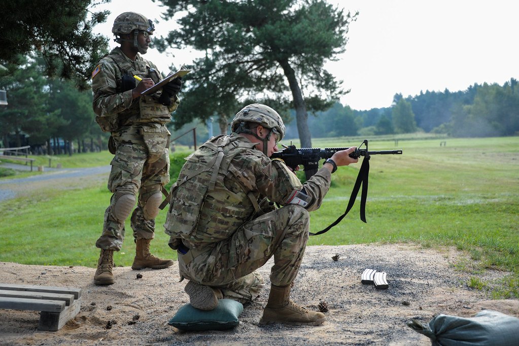 A kneeling shooter aiming a rifle on a grass range while an instructor stands behind with a clipboard, the sling hanging loose below the rifle