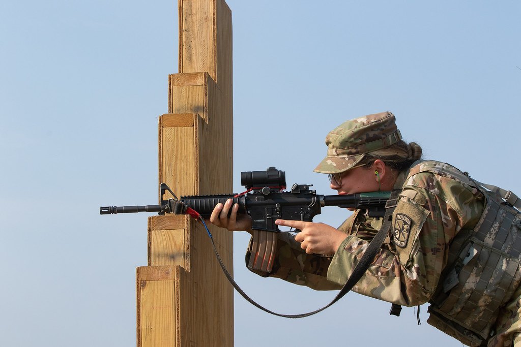 A soldier shooting a rifle around the side of a wooden barricade, with a sling attached and looping below the handguard