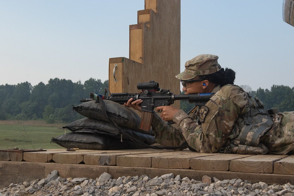 A soldier lying prone on a wooden shooting platform, resting a rifle with a magnified optic on sandbags