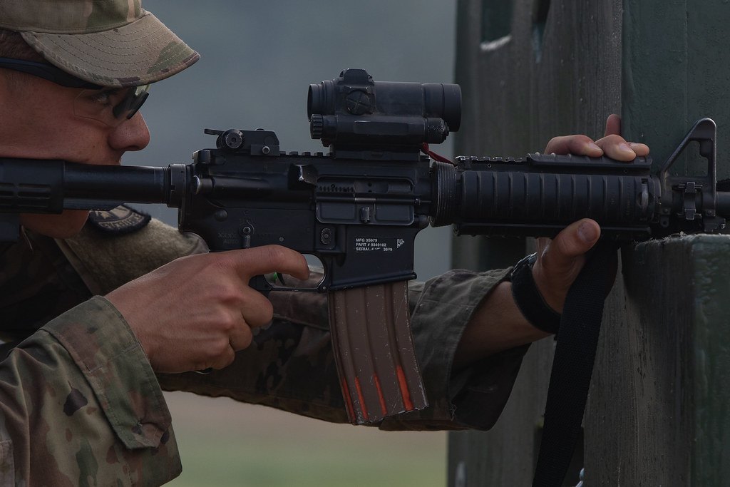 Close-up of a rifle receiver, magazine and mounted optic as a shooter fires from behind a wooden barricade