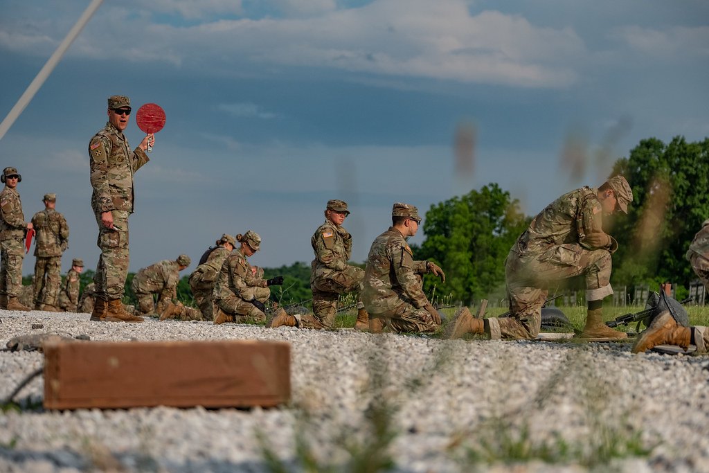 A line of soldiers kneeling and standing at firing points on a gravel range, with an instructor holding a red paddle