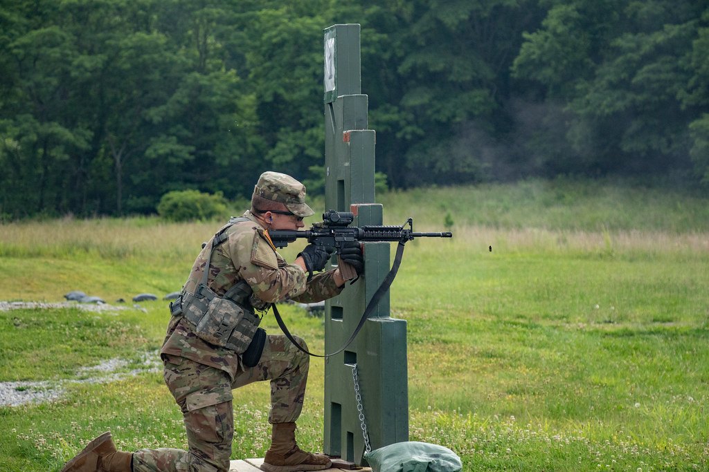 A soldier shooting a rifle from a kneeling position beside a green support post on a grass range