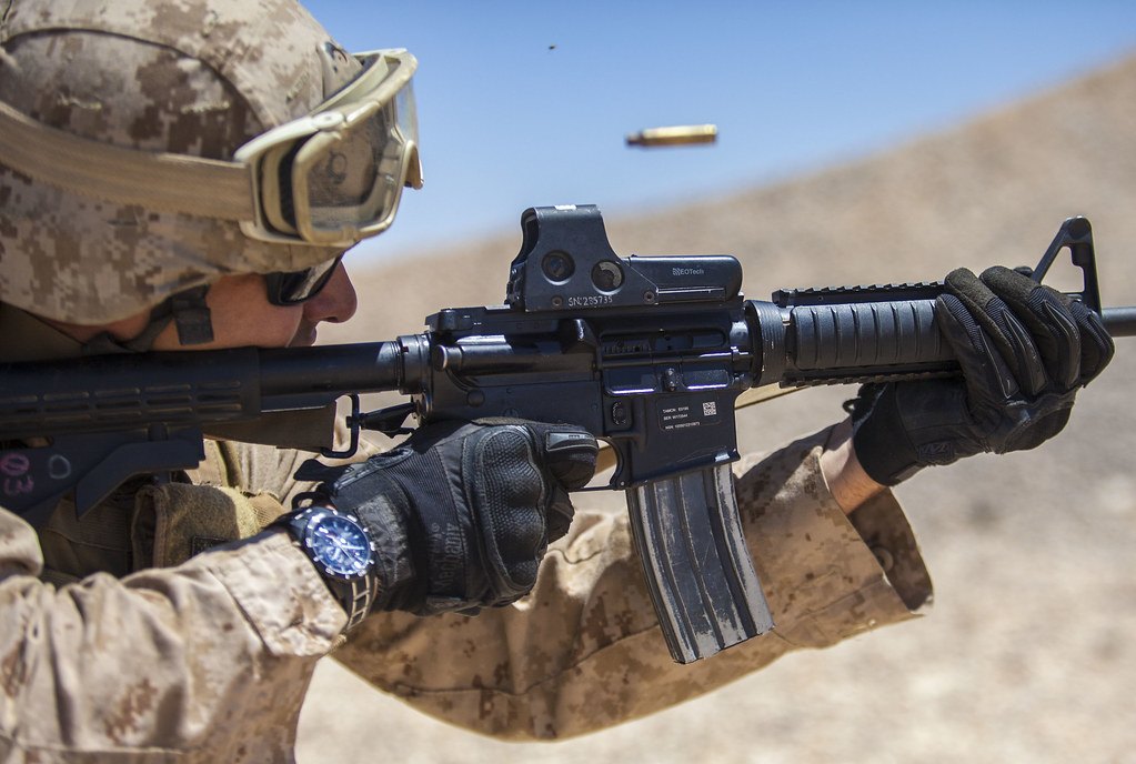 Close-up of a shooter firing a rifle fitted with a holographic sight, a spent case in the air above the ejection port