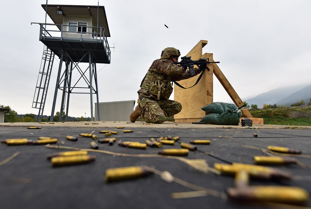 Spent brass cartridge cases in the foreground of a range, with a kneeling shooter and a range control tower behind