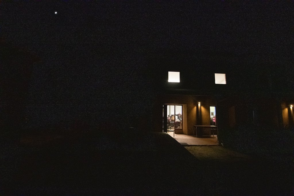 A building at night seen from outside, with light spilling from an open doorway and two upstairs windows