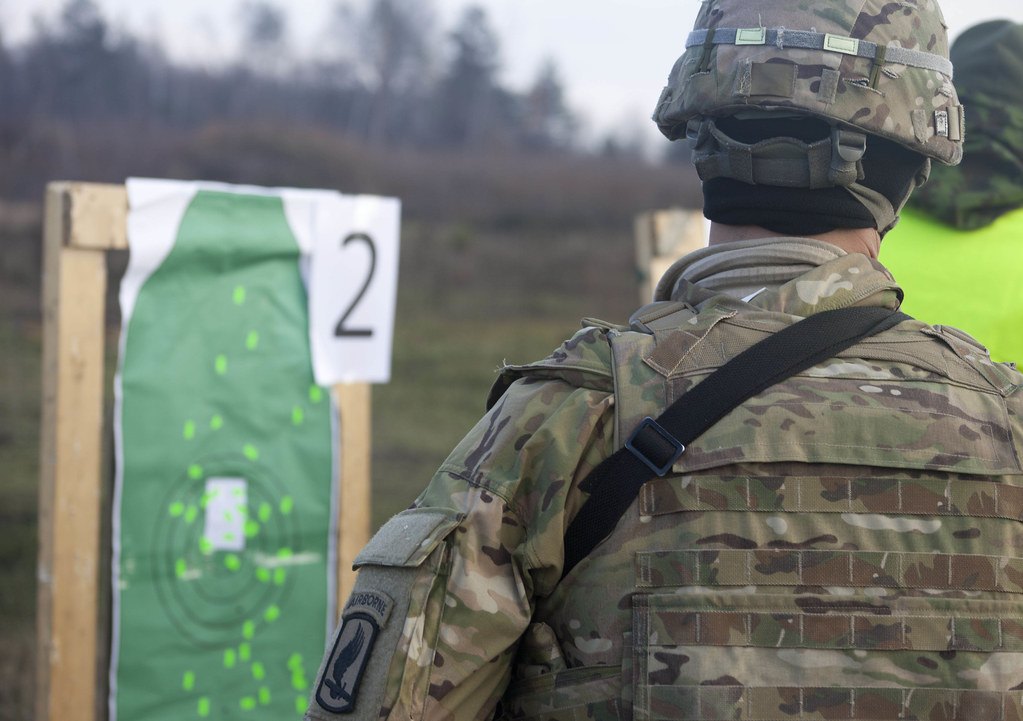 A soldier seen from behind facing a green zeroing target marked with concentric rings and clusters of shot markers, beside a numbered lane board