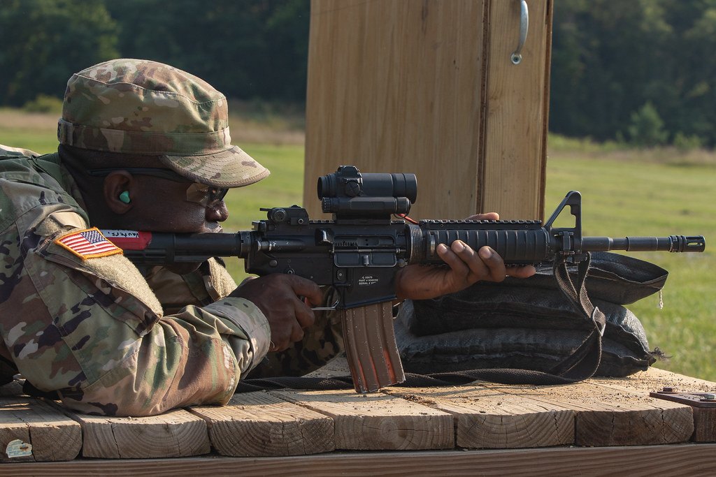 A shooter lying prone at a wooden shooting bench, firing a rifle fitted with an optic and rested on a sandbag