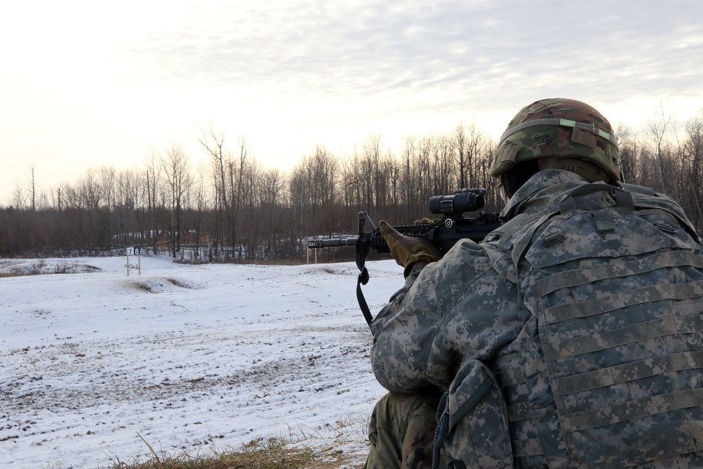 A shooter aiming a scoped rifle across a snow-covered field range under a pale sky