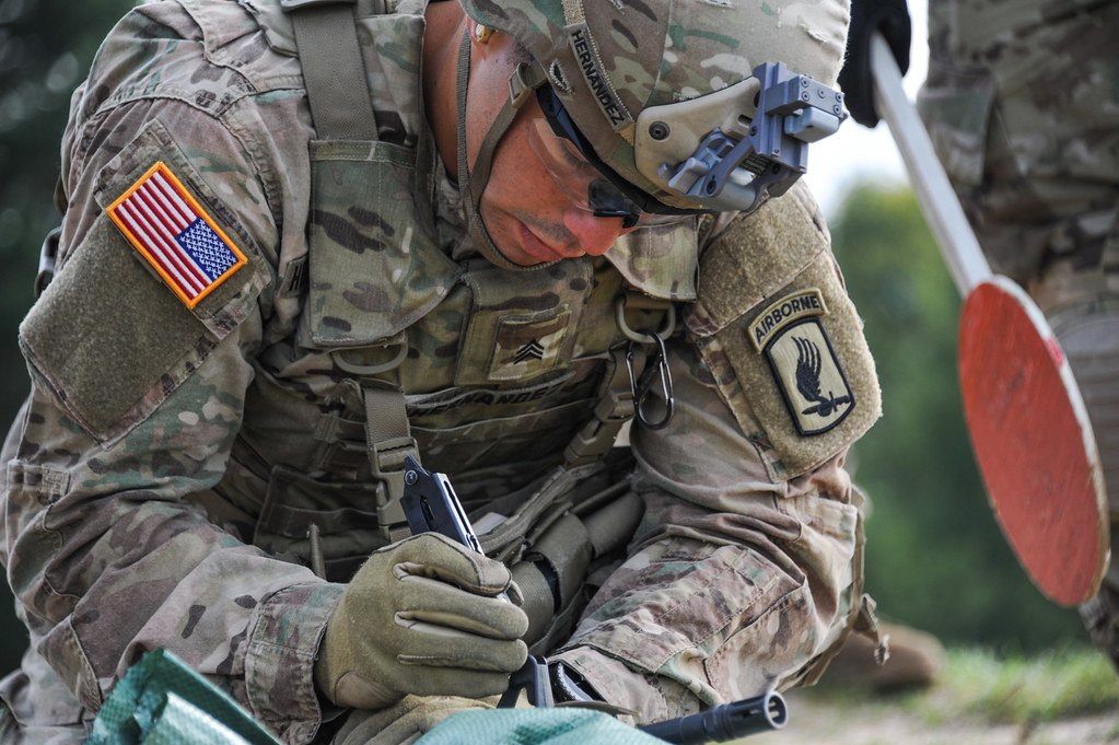 A soldier kneeling over a paper target with a pen and a red marking paddle