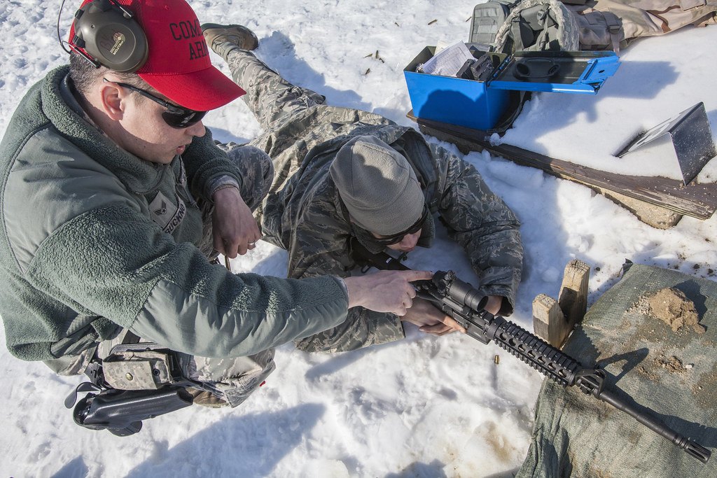 A coach leaning over a shooter on a snowy range, both handling a rifle fitted with a scope