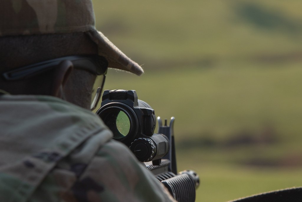 Close-up of a small optic mounted on the top rail of a rifle, with a shooter behind it