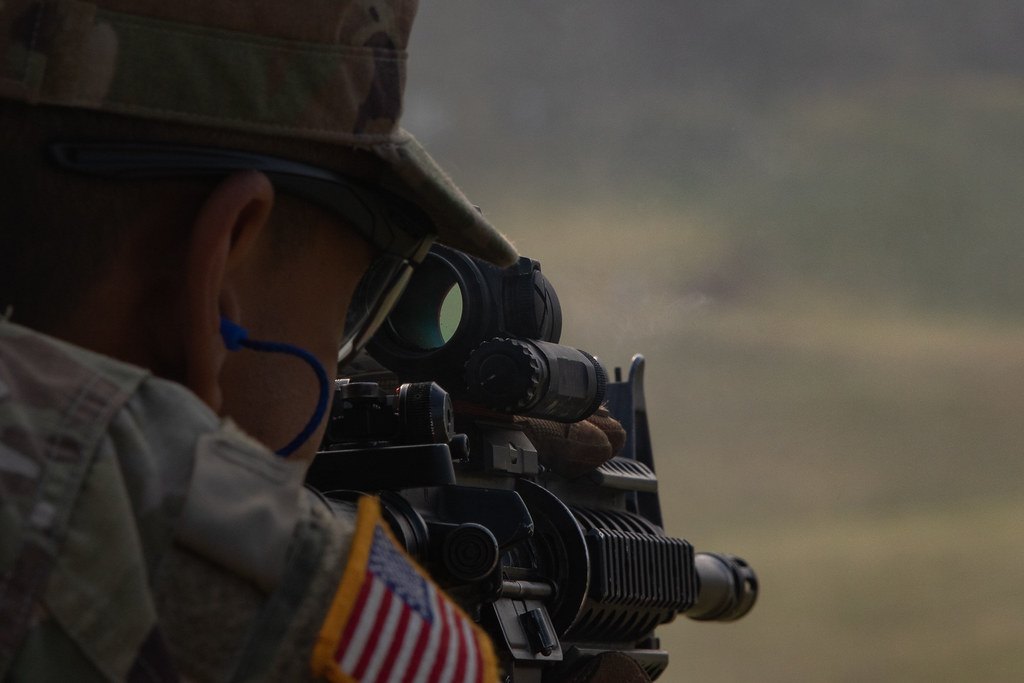 A shooter aiming a rifle fitted with an optic, seen from behind the shoulder