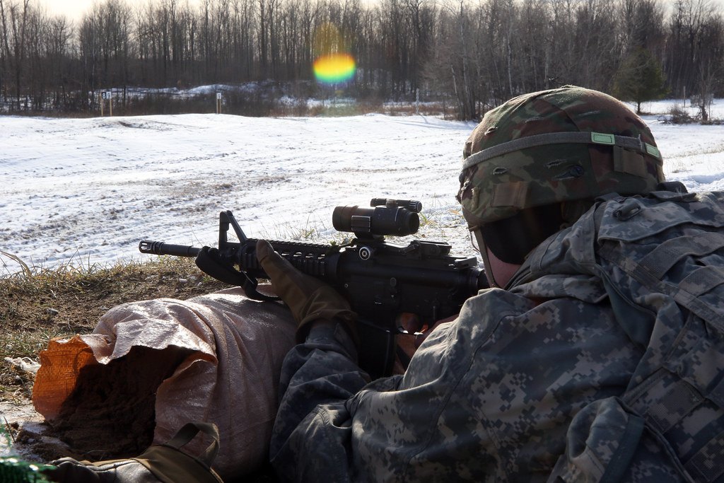 A shooter lying prone on a snow-patched range aiming a rifle fitted with a tube style red dot sight