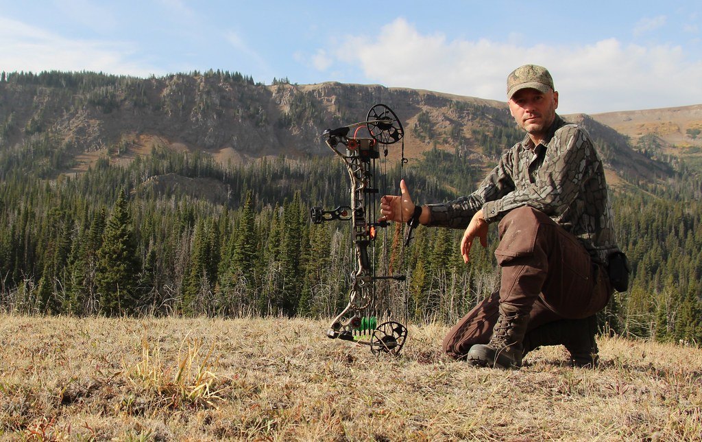 A kneeling hunter in camouflage steadying a compound bow fitted with a sight, stabiliser and quiver, mountain timber behind