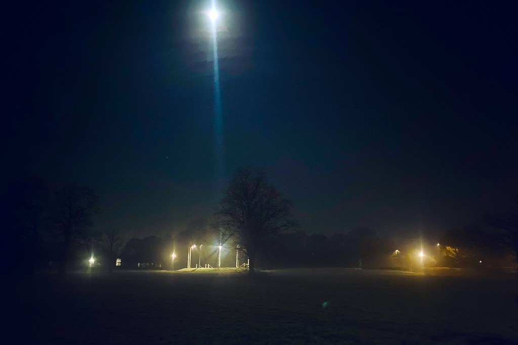 A bright light glowing above a dark misty field at night, with scattered lamps along a distant treeline