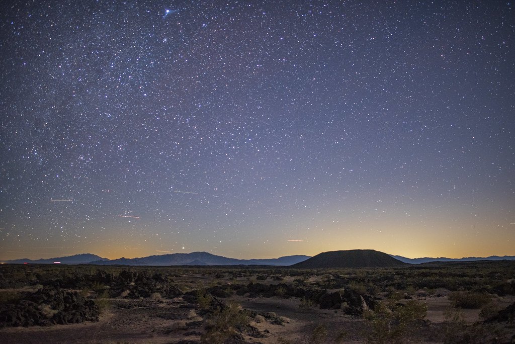 A dark desert landscape under a star filled sky with a low volcanic crater on the horizon