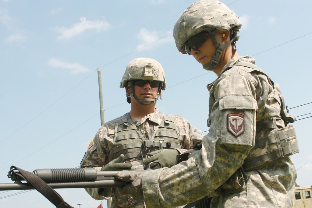Two soldiers handling a pump shotgun with a black webbing sling attached near the magazine tube