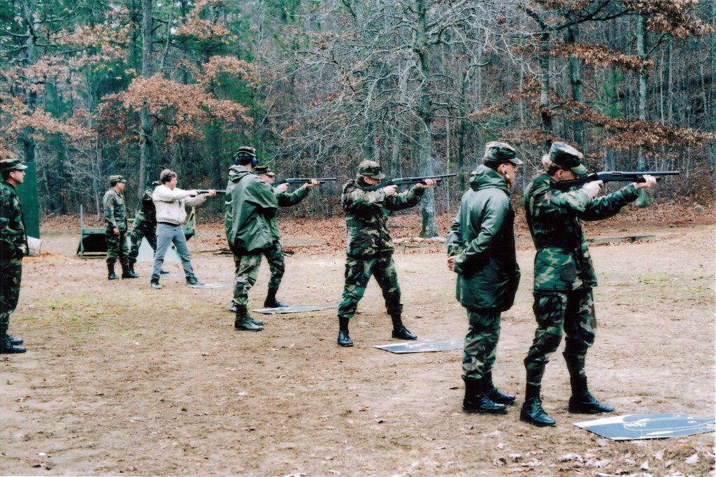 A line of shooters on a wooded outdoor range firing pump action shotguns under supervision