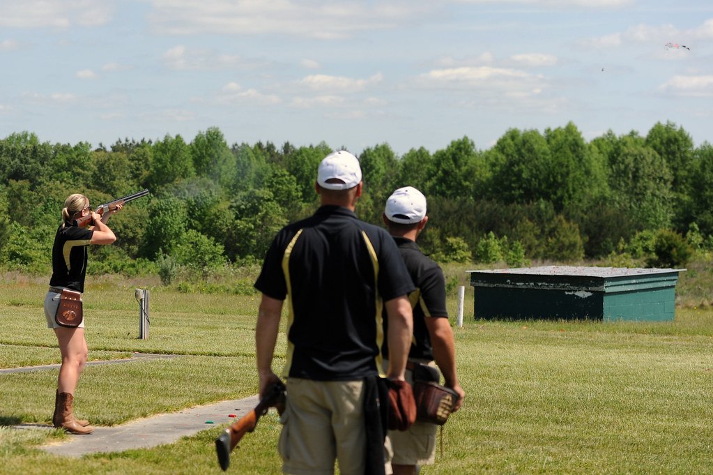 A shooter on a grass trap range taking a shot while two others wait behind with open guns