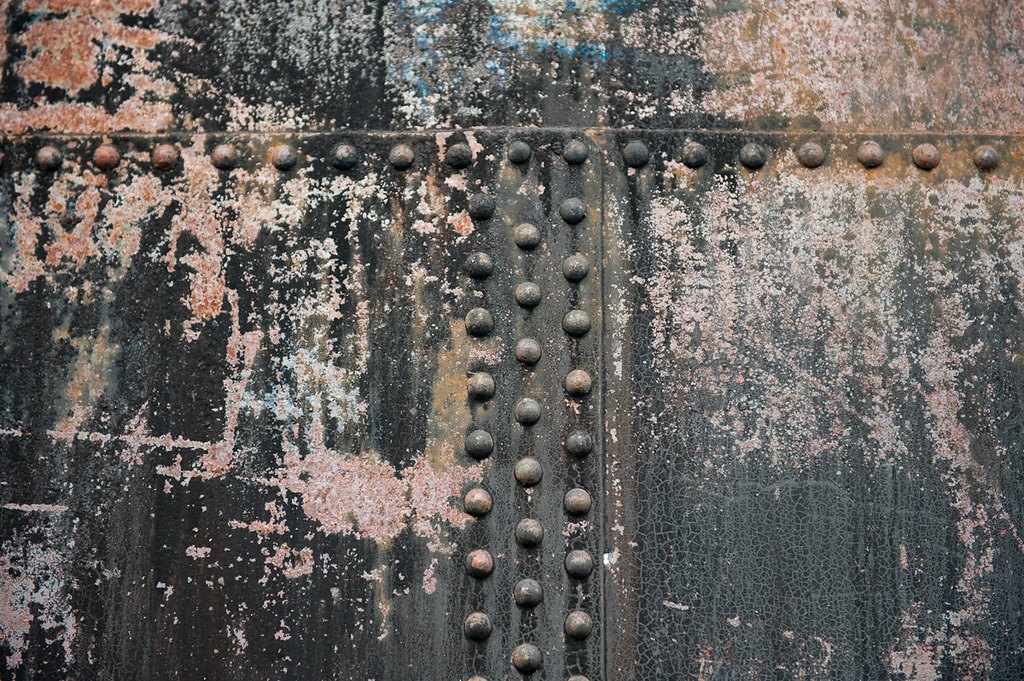 A heavily corroded riveted steel plate with paint flaking away from the rivet heads