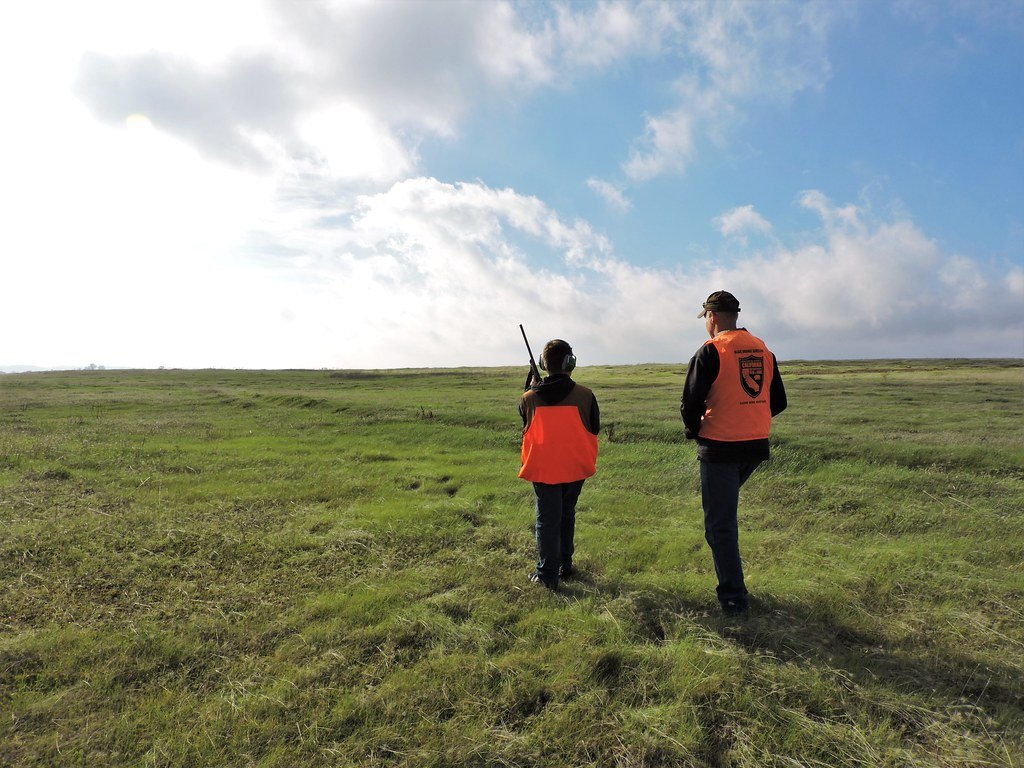 An adult and a smaller young hunter seen from behind walking away across open green grassland, the younger one carrying a shotgun muzzle up