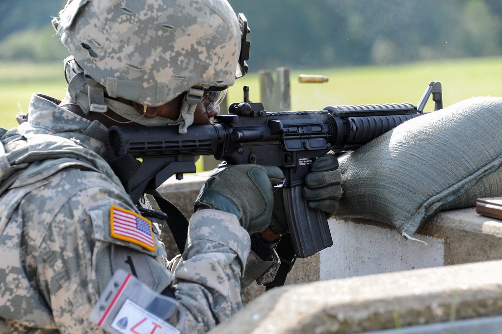 A soldier lying prone behind sandbags on a concrete firing point, aiming a rifle across a grass field