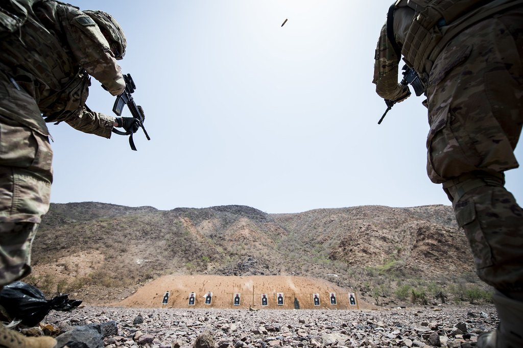 Shooters standing at a firing line on a dirt range with a row of silhouette targets against a bare hillside