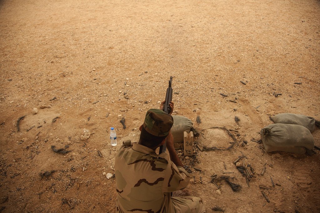 A shooter lying prone on bare desert ground behind sandbags, aiming down a dirt range