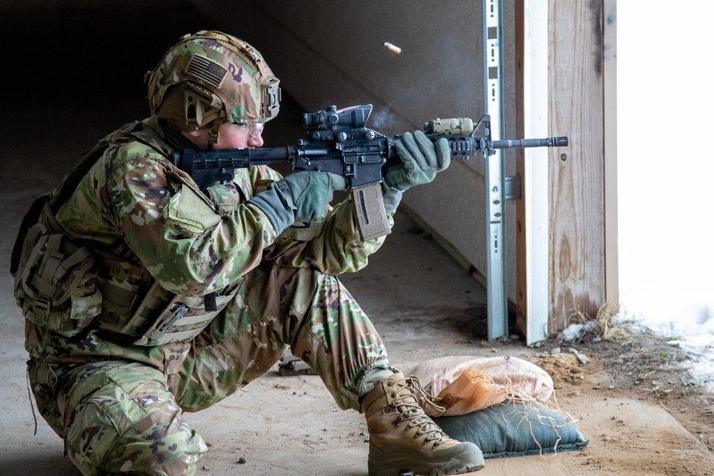 A soldier firing a rifle from a kneeling position inside a doorway, with sandbags at his feet
