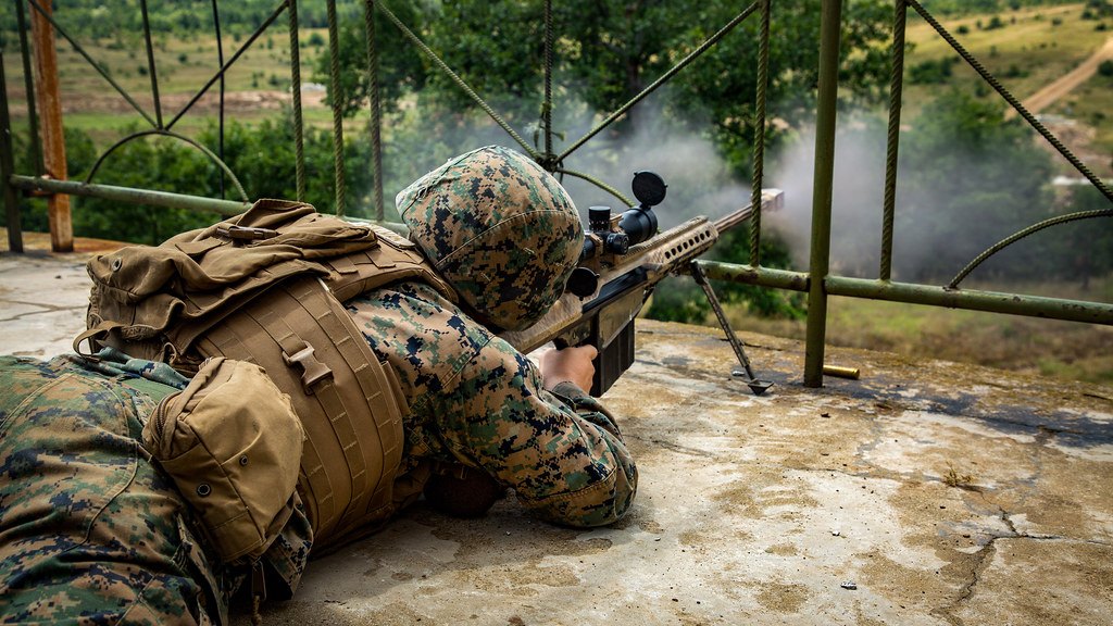 A prone shooter firing a rifle from a bipod on a range, with smoke drifting from the muzzle
