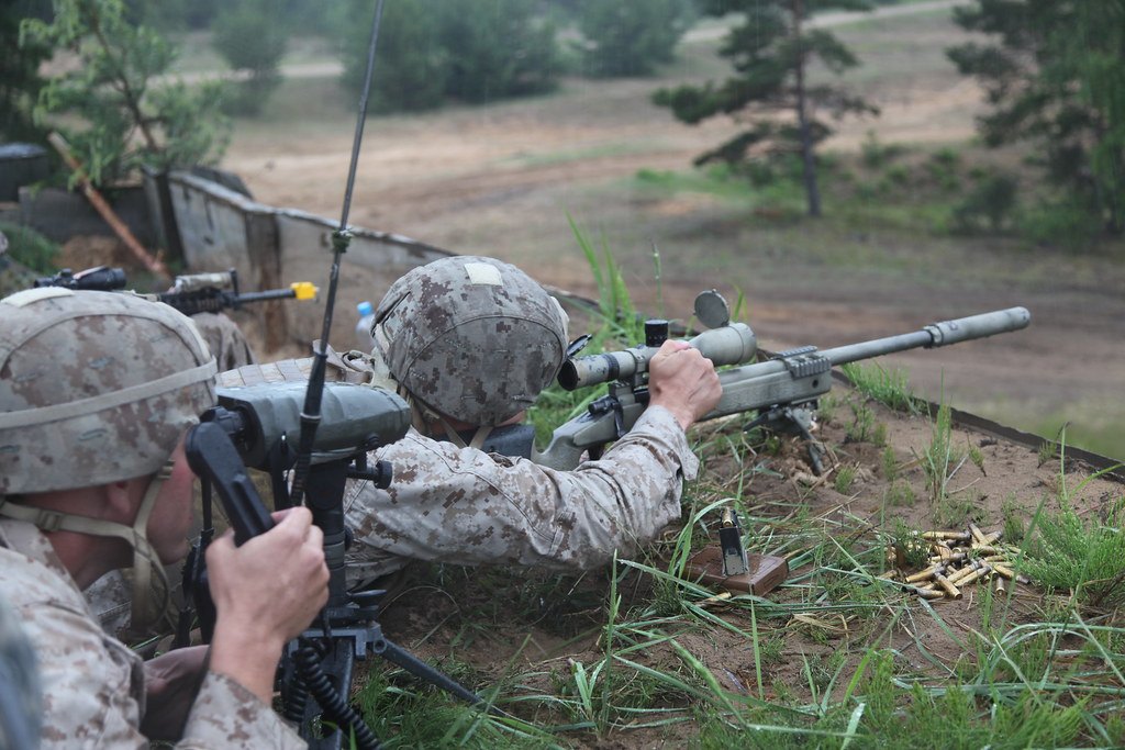 Two shooters lying prone in the grass on a range, one behind a rifle and one behind a spotting scope