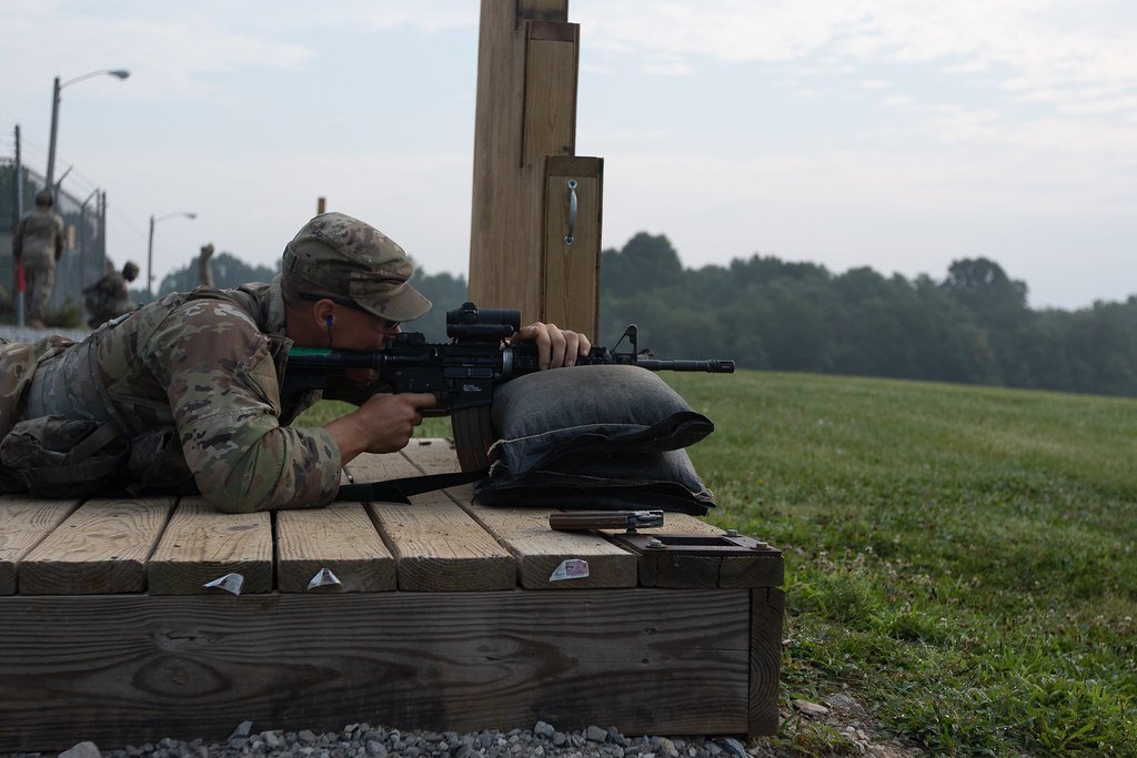 A shooter lying prone on a wooden platform behind sandbags, firing across an open green field