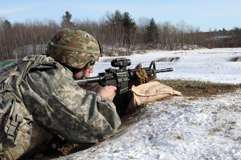 A shooter lying prone in snow-patched grass, aiming a rifle fitted with an optic